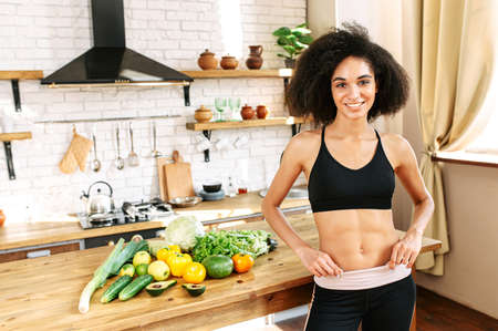 Athletic body thanks to proper nutrition. Young african-american girl with a perfect body in sportswear stands in the kitchen with hands on hips, vegetables on the table on the backgroundの写真素材
