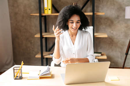 Cheerful biracial young woman uses laptop for video call. A smiling girl sits in modern office and emotionally talks via video connection. Front view. Video meeting conceptの写真素材