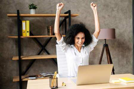 Got it! Cheerful attractive young woman with an afro hairstyle in modern office had seen good news in network, she looks at laptop screen and raised her hands up in triumph. Victory conceptの写真素材
