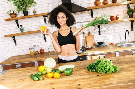 Attractive african-american woman with a athletic fit body stands in the kitchen near table full of vegetables on it, holds yellow pepper and leek in hands and looks at camera with a smileの写真素材
