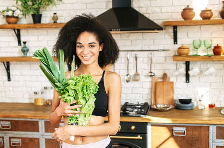 Healthy lifestyle concept. Young sporty woman in the kitchen with armful of fresh greenery and salads smilingの写真素材