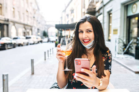 Attractive girl rests in a cafe after quarantine. A young woman with a lowered on chin protective medical mask drinks a cocktail on the terrace and uses the smartphoneの写真素材