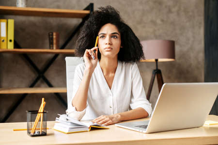Pensive mixed-race girl think about business tasks and development while sits at the desk in the office with a pencil in handの写真素材