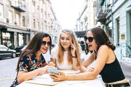 Female friends in the summer cafe on cozy european street. Three attractive young women watch into smartphone screen and talk cheerfullyの写真素材