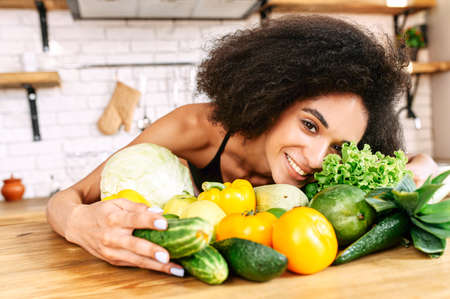 Attractive young multiethnic woman with an afro hairstyle with arms wrapped around fresh green colored vegetables and greenery looks at camera. Healthy foodの写真素材