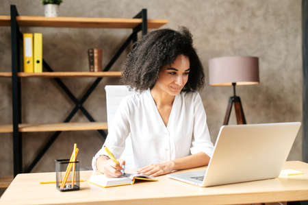 Concentrated african-american young woman in smart casual wear looks at the laptop screen while sitting in contemporary white office space and writes some notes in the notebook. Front viewの写真素材