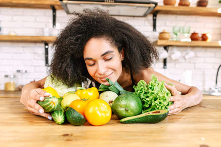 Healthy food. Close-up portrait of african-american young woman with green vegetables. A biracial girl is sniffing fresh greenery with eyes closed in the kitchenの写真素材
