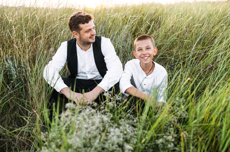 Friendship between father and preteen son. Young dad and his school-age son sits on the ground in the meadow outdoorsの写真素材