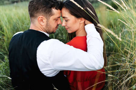 Back view of couple in love among tall grass in the meadow. A guy tenderly hugs girl, they touch their foreheadsの写真素材