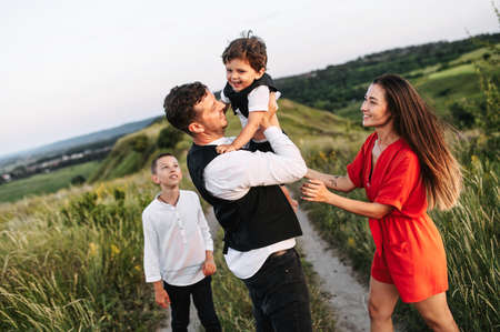 Family of four outdoors in the countryside. Young parents and their two sons - toddler and school age boy spend time together, dad carries younger son in the armsの写真素材