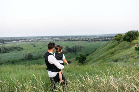 A father holds his little son in his arms with a scenic view on green hills, they look in one direction. Back viewの写真素材