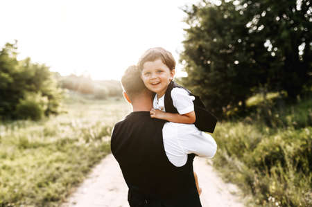 Midshot of a dad with a son on a field road. Young father stands back holding his little kid, boy is smiling to camera. Nature on the backgroundの写真素材