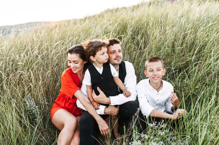 Family of four sits in a meadow in tall grass. Young parents and their two sons - toddler and school age boy. Family time conceptの写真素材