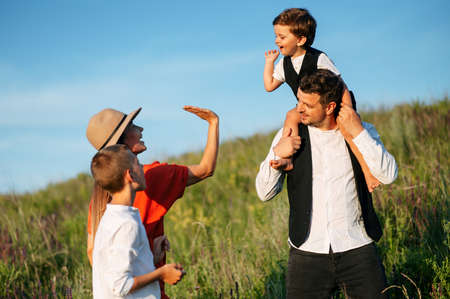 Family having fun walking on field and laughing. Girl in straw hat gives a high five to her son that sits on father's shoulder, on older happy son stands beside a motherの写真素材
