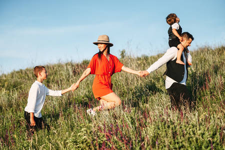Family having fun walking on field and laughing. They are holding each other by the hand. Young dad with a son sitting on shoulder and mom with the older son. Sunny day in the countrysideの写真素材