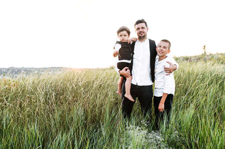 A father's love. Young dad and his two sons in the meadow outdoors, father carries toddler in his one arm and hugs preteen son with otherの写真素材