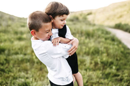 Midshot of two brothers on field hugging. Older brother holds a little one, both of them are smiling and wearing casual clothes. Green field on the backgroundの写真素材