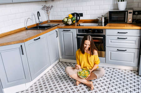Veggie lifestyle. Cheerful young woman sits on the floor in the kitchen in scandinavian style and...