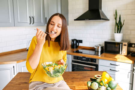 Delicious and healthy food. An attractive young girl sits on the edge of table in the kitchen and eats a fresh vitamin salad from a bowlの写真素材