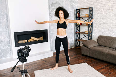 Sports vlogs. A biracial girl in sportswear does stretching, practices yoga, does fitness at home and records herself on camera in cozy atmosphere. Online coaching, trainerの写真素材