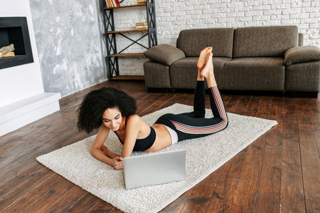 A beautiful multi-ethnic woman in sportswear is watching workout videos on a laptop at home while taking a break between sets, she lays on the floor in cozy living roomの写真素材