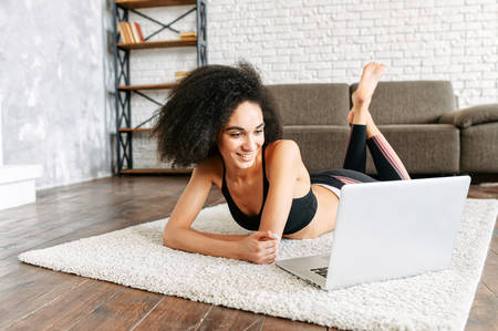 An attractive African-American girl in sportswear is watching workout videos on a laptop at home while taking a break between sets, she lays on the floor in cozy living roomの写真素材