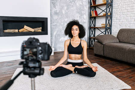 Young sporty slim mixed-race woman yoga coach records video online training yoga. An african-American girl sits in lotus pose and looks at cameraの写真素材
