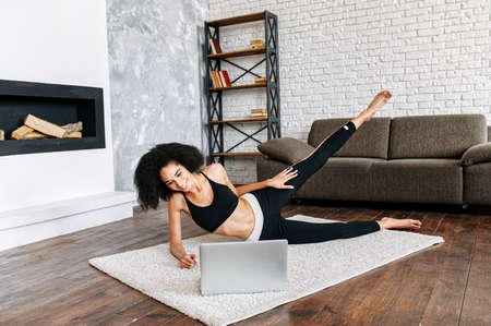 A young African-American woman is doing sports on video lessons. The girl in sportswear is watching stretch tutorials on laptop at home and repeating exercisesの写真素材