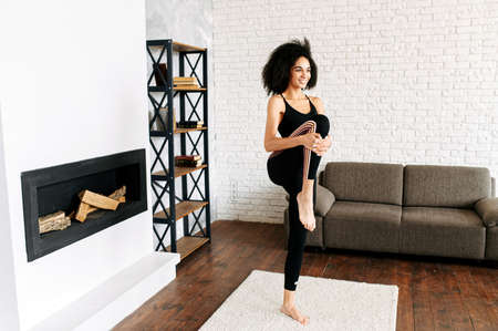 Warm up before training at home. An African-American beautiful girl in sportswear is doing stretching legs in cozy living roomの写真素材