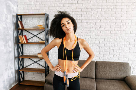 A beautiful African-American woman in sportswear is doing workout at home, the girl stands with skipping rope on the shoulders and looks at cameraの写真素材