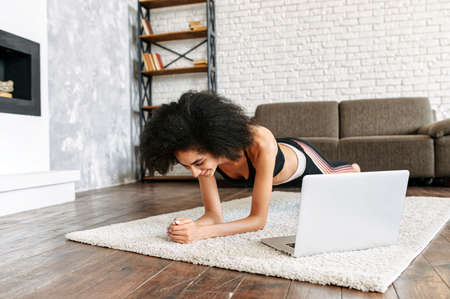Get in shape at home. A young biracial girl uses a workout video tutorial, she sits on a mat in the room, does exercises and looks at the laptop. Selfdissipline and healthy lifestyle conceptの写真素材