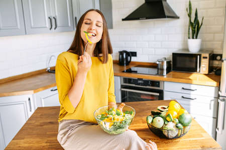 Delicious and healthy food. An attractive young girl sits on the edge of table in the kitchen and eats a fresh vitamin salad from a bowlの写真素材