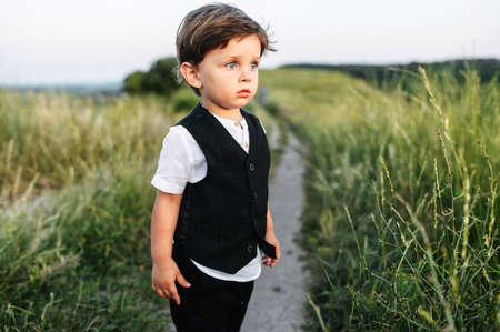Toddler with a big blue eyes in white shirt and suit vest stands and looks away in the countryside. Cute little boy on the dirt road through meadowの写真素材
