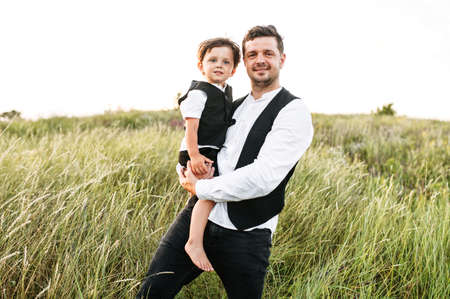 A toddler boy in arms of his father outdoors. Father and little son in suit vests walk on the meadow, they look at cameraの写真素材