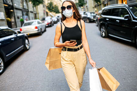 Young woman has a shopping day after relief quarantine period. Beautiful girl in protective medical mask is walking with shopping packages in the cityの写真素材