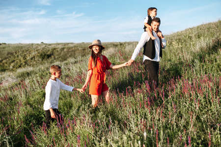 Happy family walking through the field of flowers. The first one is a father with a boy on his shoulders, he holds a wife's hand that walks behind. She holds an older son, they are kindly smilingの写真素材