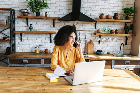 Smiling beautiful young woman speaks on the phone while sitting at the kitchen table In front of the laptop and notebook. She works from home on a cozy wooden kitchen.の写真素材