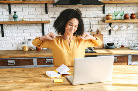Smiling good looking afro-american girl stretches while sitting at the kitchen table with a laptop and notebook on it. Working from home, wooden kitchenの写真素材