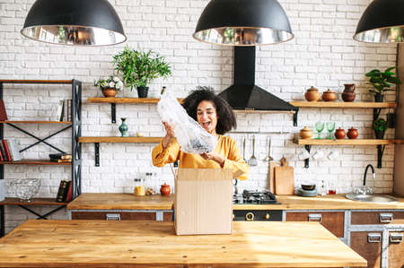 Unpacking the long-awaited package. A young African-American girl excitedly takes out an internet order from a box standing in a cozy kitchen at homeの写真素材