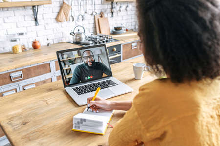 A female student studies online. An african woman is watching online classes and writing a syllabus in notebook, black male tutor on the screen. Concept of distance study, online learning, webinarsの写真素材