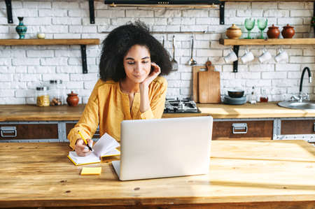 Smiling young girl with an afro hair style is studying at the kitchen table using a laptop. She is leaning on a chin, making notes in her notebook. Front midshotの写真素材