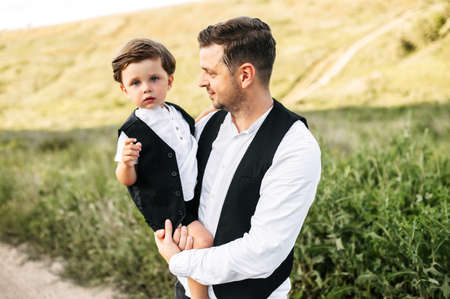 Side shot of a young man with his son. Young attractive man holds a kid on his hands and looking at him. A little kid looks like his father points with a finger to a camera. Nature is on the backgroundの写真素材