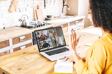 Morning meeting online. A young african-american woman is using app on pc for connection with colleagues, employees. Video call with many people together. Back viewの写真素材