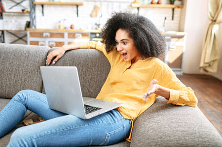 Unexpected news. Surprised multi-ethnic girl lies on a comfortable sofa with a laptop and looks at the screen uncomprehendingの写真素材