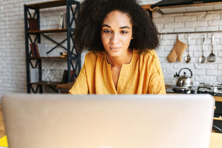 Close-up portrait of concentrated african woman uses laptop for work from home. A girl with an afro hairstyle focused on screenの写真素材