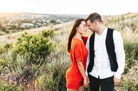 Love story shot of a young beautiful couple with nature on the background. A girl in red short overalls leans on her boyfriend. Romantic couple in love standing on the field roadの写真素材