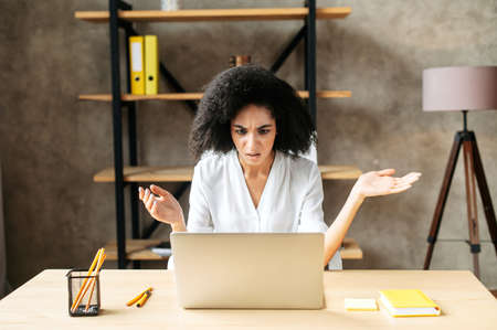 Misunderstanding at work. A puzzled african-american woman looks at the laptop screen and shrugsの写真素材