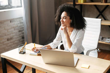 Pensive young african-american woman with a laptop in the modern office. Thoughtful girl looks throw the window and waiting for inspiration and new ideasの写真素材