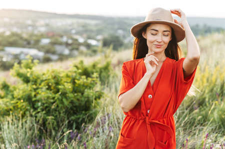 Romantic long haired girl in a hat is posing in the rural landscape, woman in red romper with a scenic view on the backgroundの写真素材