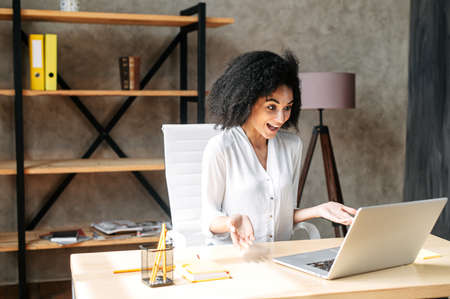 Video meeting in the office. Happy african-american woman glad to see employee or colleague via video call, she uses laptop and looks at screen cheerfully. Front viewの写真素材